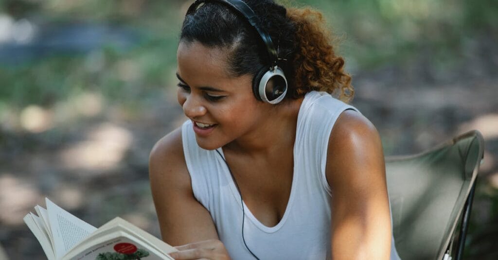 Cheerful young Hispanic female in white tank top listening to music via headphones and reading book while sitting on chair in park